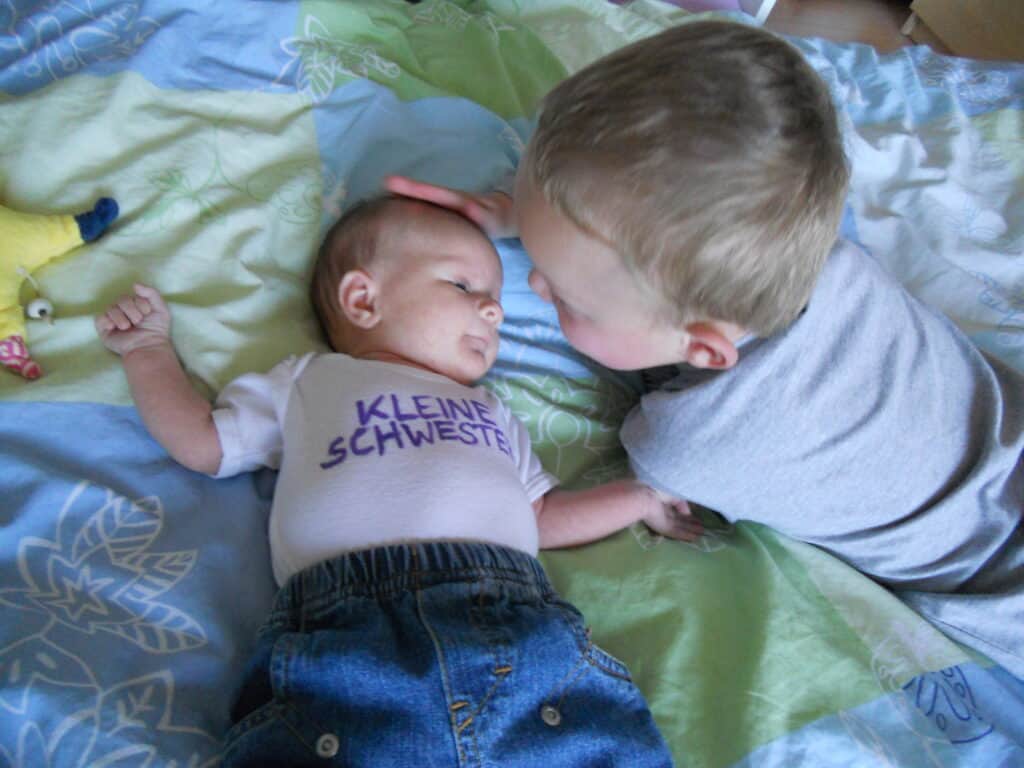 Ein Baby trägt ein Shirt mit der Aufschrift Kleine Schwester. Ein älteres Kind streichelt das Baby am Kopf. Das ist das Bonuskind, der Halbbruder.